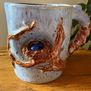 May include: A ceramic mug with a light blue speckled glaze. The mug features a sculpted bird's nest with two blue eggs and brown branches wrapping around the body and handle. The handle is also textured to resemble a branch.