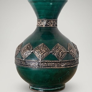May include: A deep emerald green ceramic vase with a flared neck and a rounded body. The vase features metallic silver-toned decorative bands around the neck and midsection, with a repeating pattern of embossed diamond shapes and intricate scrollwork.