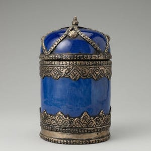 May include: A cylindrical blue container with a domed lid, featuring intricate silver-toned metal filigree bands around the base, the middle, and the lid. The metalwork displays detailed engraved patterns, and the lid is topped with a small finial.