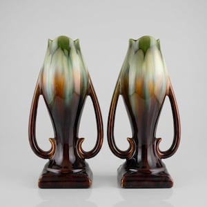 Peut inclure: Deux vases en céramique au design unique. Les vases ont une base et un corps brun foncé, avec un glaçage brun clair et vert sur la partie supérieure. Chaque vase présente des anses incurvées et une base carrée.
