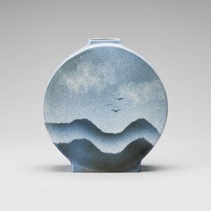May include: A circular ceramic vase featuring a blue and white speckled glaze. The surface displays a painted landscape of layered dark blue mountain silhouettes against a lighter blue sky with soft clouds and two small black birds in flight.
