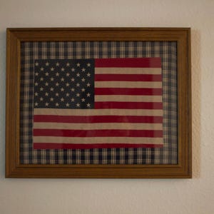Peut inclure: Œuvre d'art encadrée du drapeau américain. Le drapeau présente des rayures rouges et blanches avec un champ bleu d'étoiles blanches. Il est encadré d'un cadre en bois marron et d'une bordure en tissu à carreaux.
