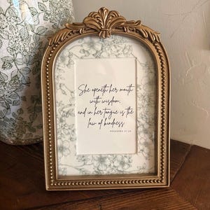 May include: An arched gold picture frame with a beaded border and a decorative top crest. Inside, a white card features a gray floral pattern and black text that reads: She openeth her mouth with wisdom; and in her tongue is the law of kindness. Proverbs 31:26.