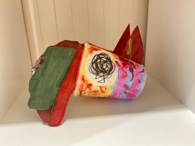 May include: A handcrafted sculpture made from a paper cup and cardboard. The cup features a white circle with a black scribbled design, surrounded by red, yellow, and blue paint. A green and red cardboard piece is attached to the side of the cup.