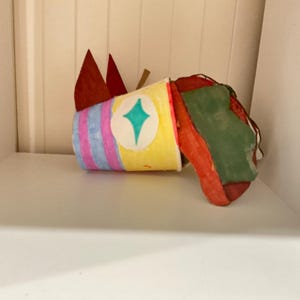 May include: A handcrafted paper cup sculpture featuring a yellow body with vertical pink and blue stripes, a central teal diamond shape, and two pointed red paper ears. Attached to the side is a textured, irregularly shaped red and green form.
