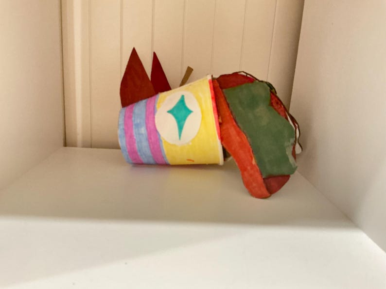 May include: A handcrafted paper cup sculpture featuring a yellow body with vertical pink and blue stripes, a central teal diamond shape, and two pointed red paper ears. Attached to the side is a textured, irregularly shaped red and green form.