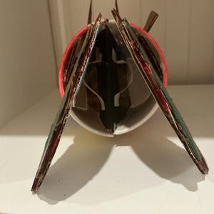 May include: A cylindrical white cup structure featuring two angled cardboard panels attached to the sides. The panels are painted dark green with red edges. A red circular rim is visible at the top of the white cup, with internal white supports.