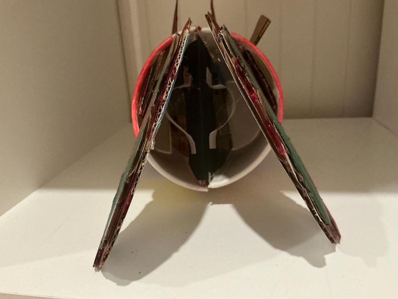 May include: A cylindrical white cup structure featuring two angled cardboard panels attached to the sides. The panels are painted dark green with red edges. A red circular rim is visible at the top of the white cup, with internal white supports.