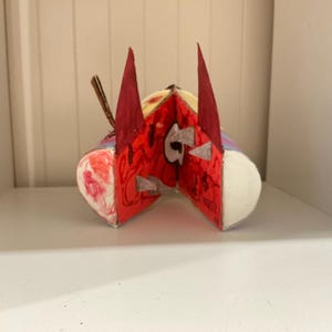 May include: A handcrafted sculpture featuring a central red face with two tall, pointed dark red ears. The figure has a wide, open mouth revealing white teeth, with additional red and white painted details on the sides and a small brown stick element.
