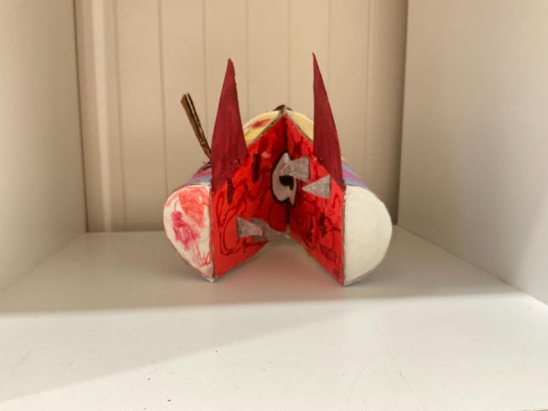 May include: A handcrafted sculpture featuring a central red face with two tall, pointed dark red ears. The figure has a wide, open mouth revealing white teeth, with additional red and white painted details on the sides and a small brown stick element.