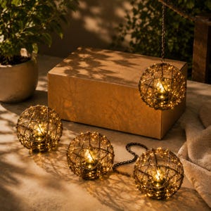 May include: Decorative spherical lights with warm white LED lights encased in a woven metal frame. The lights are arranged in a cluster of four, with one hanging from a chain and the others resting on a surface. A cardboard box is in the background.