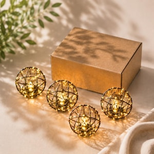 May include: Four decorative, spherical lights with warm white LED lights encased in a bronze wire frame. A brown cardboard box sits behind the lights. The lights are on a light-colored surface with shadows.