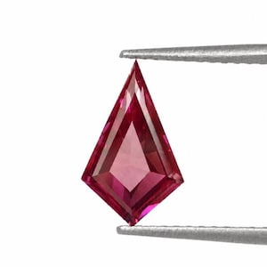 Puede incluir: Una gema facetada en forma de cometa, en tonos rosas y rojos, es sostenida por unas pinzas plateadas sobre un fondo blanco. La gema tiene un diseño simétrico con ángulos agudos y una superficie pulida que refleja la luz.