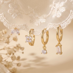 Puede incluir: Un conjunto de cuatro pendientes de aro dorados con piedras transparentes facetadas. Incluye un diseño con dos piedras en talla marquesa, una flor de cuatro pétalos, un colgante solitario redondo y un colgante rectangular en talla baguette.