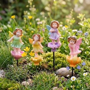 May include: Four colorful fairy garden stakes. Each fairy has a different colored dress and wings, with a flower crown. The fairies are standing on a flower base, with a black stake. The background is a garden with green grass and flowers.