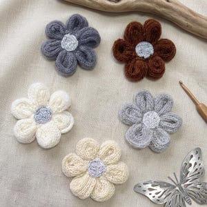 Könnte beinhalten: Fünf handgefertigte, gestrickte Blumenapplikationen in verschiedenen Farben: Grau, Braun, Creme und Off-White. Jede Blume hat eine silberne Mitte. Ein Häkelhaken und ein Metallschmetterling sind ebenfalls sichtbar.