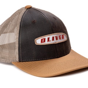 May include: A black and brown trucker hat with a tan brim. The hat has a white patch with the word "OLIVER" in red embroidered letters.