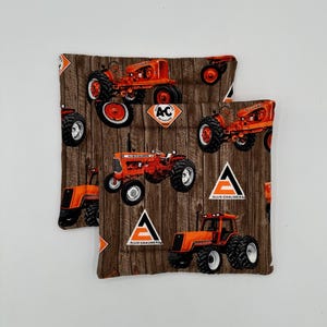 Puede incluir: Dos cuadrados de tela con un patrón de vetas de madera marrón. La tela está estampada con tractores naranjas y el logotipo de Allis-Chalmers. Los tractores tienen neumáticos negros y están en varias posiciones.