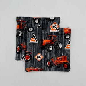 Puede incluir: Dos posavasos de tela gris y naranja con un patrón de veta de madera. Los posavasos presentan un patrón repetido de tractores naranjas con el texto "Allis-Chalmers" y "AC" en ellos.
