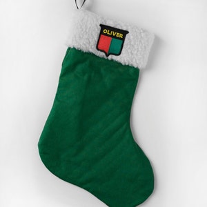 May include: A green Christmas stocking with a white, fluffy cuff. The stocking has a black loop for hanging and a patch with the name "OLIVER" above a red and green crest. A festive holiday decoration.