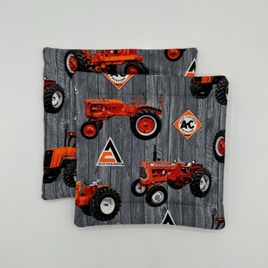 Puede incluir: Dos posavasos de tela gris y blanca con un patrón de veta de madera. Los posavasos presentan un patrón repetido de tractores rojos y naranjas con el logotipo de Allis-Chalmers.
