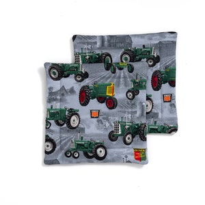 Puede incluir: Dos artículos cuadrados de tela con un fondo gris que presentan tractores verdes con detalles en rojo y amarillo. Los tractores están en varias posiciones, y la tela tiene un diseño de escena de granja. Un artículo es ligeramente más pequeño que el otro.