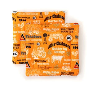 Puede incluir: Dos cojines cuadrados de tela naranja con un patrón repetido de ilustraciones de tractores blancos, texto y logotipos. El texto incluye "Allis-Chalmers", "Tractors" y "Snap-Coupler". Los cojines son probablemente para la decoración del hogar o manualidades.