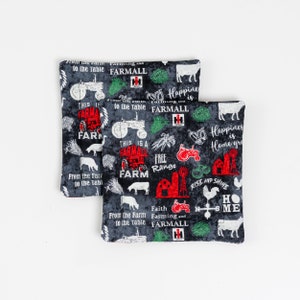 Puede incluir: Dos artículos cuadrados de tela con temática de granja con un fondo gris oscuro. La tela está estampada con gráficos y texto en rojo, verde y blanco relacionados con la granja, incluyendo tractores, vacas y las palabras "Granja" y "Felicidad".