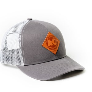 May include: A gray and white trucker hat with a brown leather patch featuring the text "Allis Chalmers" and the logo "AC".