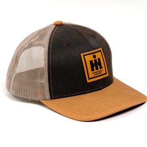May include: A brown and tan trucker hat with a mesh back. The front panel is dark brown with a tan brim. A square patch with the words "INTERNATIONAL HARVESTER" is on the front.