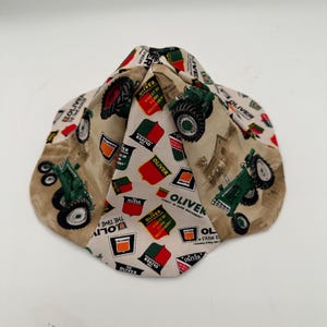 May include: A beige fabric hat featuring a repeating pattern of green tractors, red and green shields, and the word "OLIVER" in various fonts. The hat has a unique, folded design.