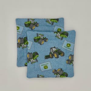 May include: Two square, light blue fabric items with a pattern of green and yellow tractors and the John Deere logo. The tractors are in various positions, and the fabric has a denim-like texture.