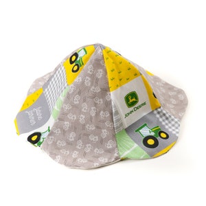 May include: A gray and yellow fabric hat with a John Deere logo and tractor designs. The hat has a patchwork design with a gray and white checkered pattern and a yellow and green pattern.