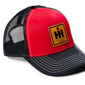 May include: Red and black trucker hat with a curved brim and mesh back. The front panel is red, and the brim is black with white stitching. A square patch with the International Harvester logo is on the front.