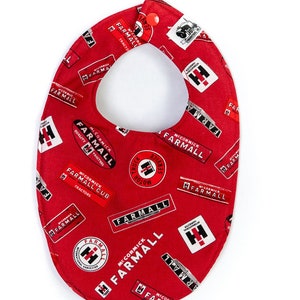 May include: Red fabric baby bib with a white and black pattern of the Farmall tractor logo. The bib has a snap closure at the neck.