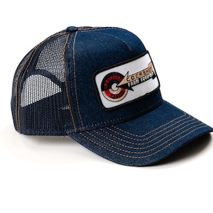 May include: A blue denim trucker hat with a mesh back. The hat features a rectangular patch with the text "COCKSHUT FARM EQUIPMENT" and a circular logo. The hat has a curved brim and is stitched with tan thread.