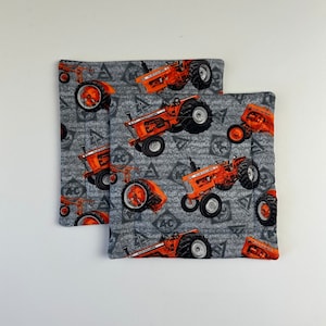 Puede incluir: Dos agarraderas cuadradas acolchadas con un fondo gris y un patrón repetido de tractores naranjas. Los tractores tienen neumáticos negros y están etiquetados "AC". Las agarraderas están bordeadas con una tela gris más oscura.