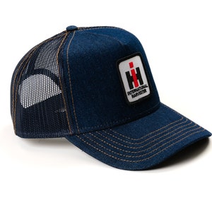 Puede incluir: Una gorra de camionero de mezclilla azul con una parte trasera de malla negra. La gorra tiene una visera cosida marrón y un parche con el logotipo de International Harvester en rojo y blanco.