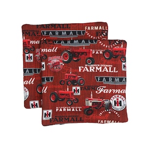 Puede incluir: Dos cojines decorativos rojos y marrones con temática de tractores Farmall. Los cojines presentan varias ilustraciones de tractores rojos y el logotipo de Farmall, junto con el texto "FARMALL" y "McCormick-Deering". Los cojines son cuadrados.