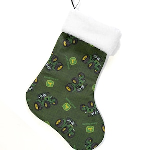 May include: Green Christmas stocking with a white faux fur cuff and a black hanging loop. The stocking is patterned with green tractors and the John Deere logo. A festive holiday decoration.