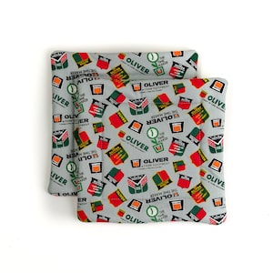 Puede incluir: Dos agarraderas de olla de tela gris y blanca con un patrón verde y rojo. El patrón presenta las palabras "Oliver" y "The Time Maker" repetidas a lo largo. Las agarraderas de olla están hechas de tela acolchada.