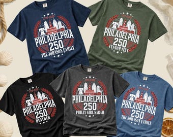 May include: Five short-sleeved t-shirts in navy, olive green, black, charcoal grey, and blue. Each shirt features a graphic with the Philadelphia skyline, the Liberty Bell, the text Philadelphia 250, and laurel wreaths in red and white ink.