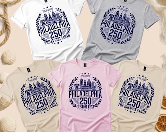 May include: Five short-sleeved t-shirts in white, grey, beige, light pink, and cream. Each shirt features a dark blue circular graphic with a Philadelphia skyline, the text 1776-2026, Philadelphia 250, and various personalised names or custom text.