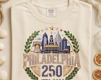 May include: A cream-coloured t-shirt featuring a graphic of the Philadelphia skyline, the Liberty Bell, and Independence Hall. The design includes the text "PHILADELPHIA 250" and "1776-2026" surrounded by a wreath of green and gold gems.