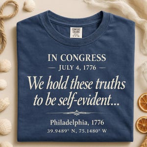 May include: A navy blue t-shirt with the text "IN CONGRESS - JULY 4, 1776 - We hold these truths to be self-evident..." and "Philadelphia, 1776" printed in white. The shirt also includes the coordinates 39.9489° N, 75.1480° W.