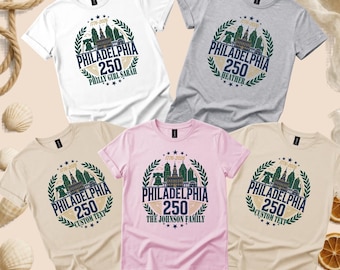May include: Five short-sleeved t-shirts in white, grey, beige, and pink. Each shirt features a circular graphic with the Philadelphia skyline, the text 1776-2026, PHILADELPHIA 250, and various custom names like Philly Girl Sarah, Heather, and The Johnson Family.