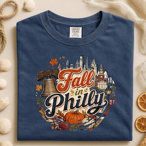 May include: A navy blue crewneck t-shirt featuring a graphic print of the Philadelphia skyline, the Liberty Bell, and autumn elements including pumpkins and leaves. The text reads Fall in Philly in a stylized orange and dark blue font.