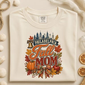 May include: A cream-colored t-shirt featuring a graphic print of the Philadelphia skyline, the Liberty Bell, a pumpkin, an apple, and autumn leaves. The text reads Philadelphia Fall MOM in a mix of script and block lettering with a plaid bow.