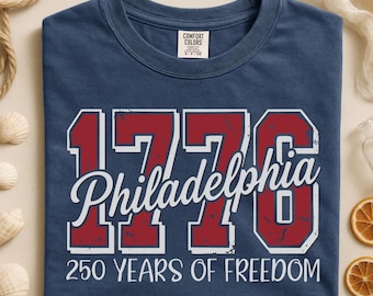 May include: Navy blue t-shirt featuring the text "1776 Philadelphia" in red and white, and "250 YEARS OF FREEDOM" in white. The shirt is a Comfort Colors brand.