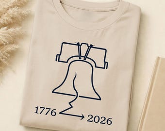 May include: Beige t-shirt with a navy blue outline of the Liberty Bell. The shirt has the numbers "1776" and "2026" with an arrow pointing from the former to the latter. The t-shirt is folded and resting on a light beige surface.